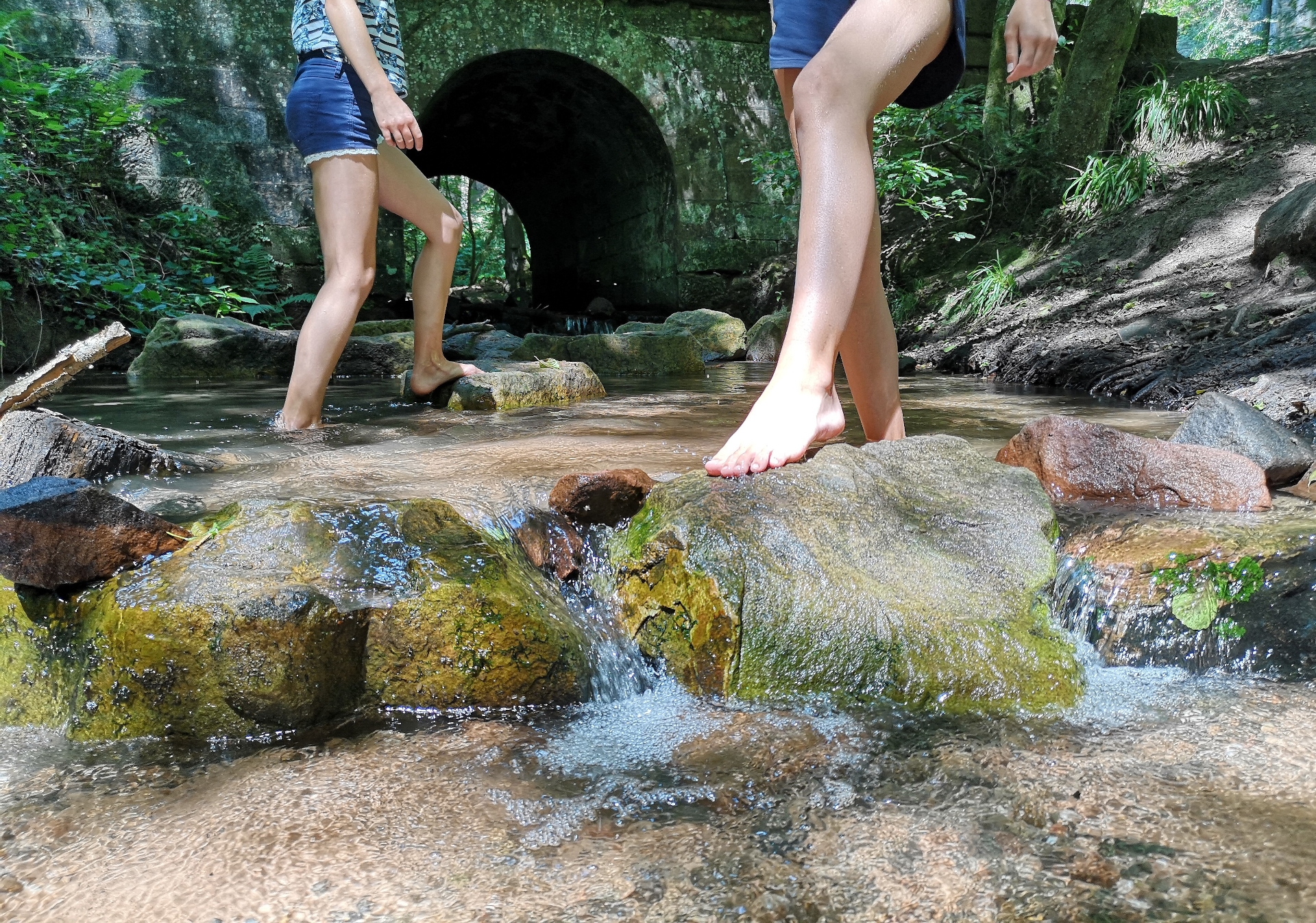 Im Vordergrund das Bachbett mit großen bemoosten Steinen. Darauf balancieren zwei barfüßige Wanderinnen.