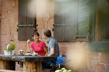 Zwei Wanderer rasten und essen auf einer Bank vor der Waldgaststätte Lindenklause. Im Rücken die rote Sandsteinmauer und zwei Fenster der Gaststätte.
