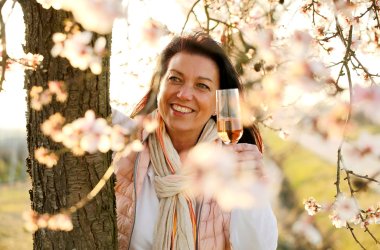 Durch die rosa blühenden Zweige eines Mandelbaums im Vordergrund, sieht man eine lächelnde Frau mit einem Glas Rose-Sekt.
