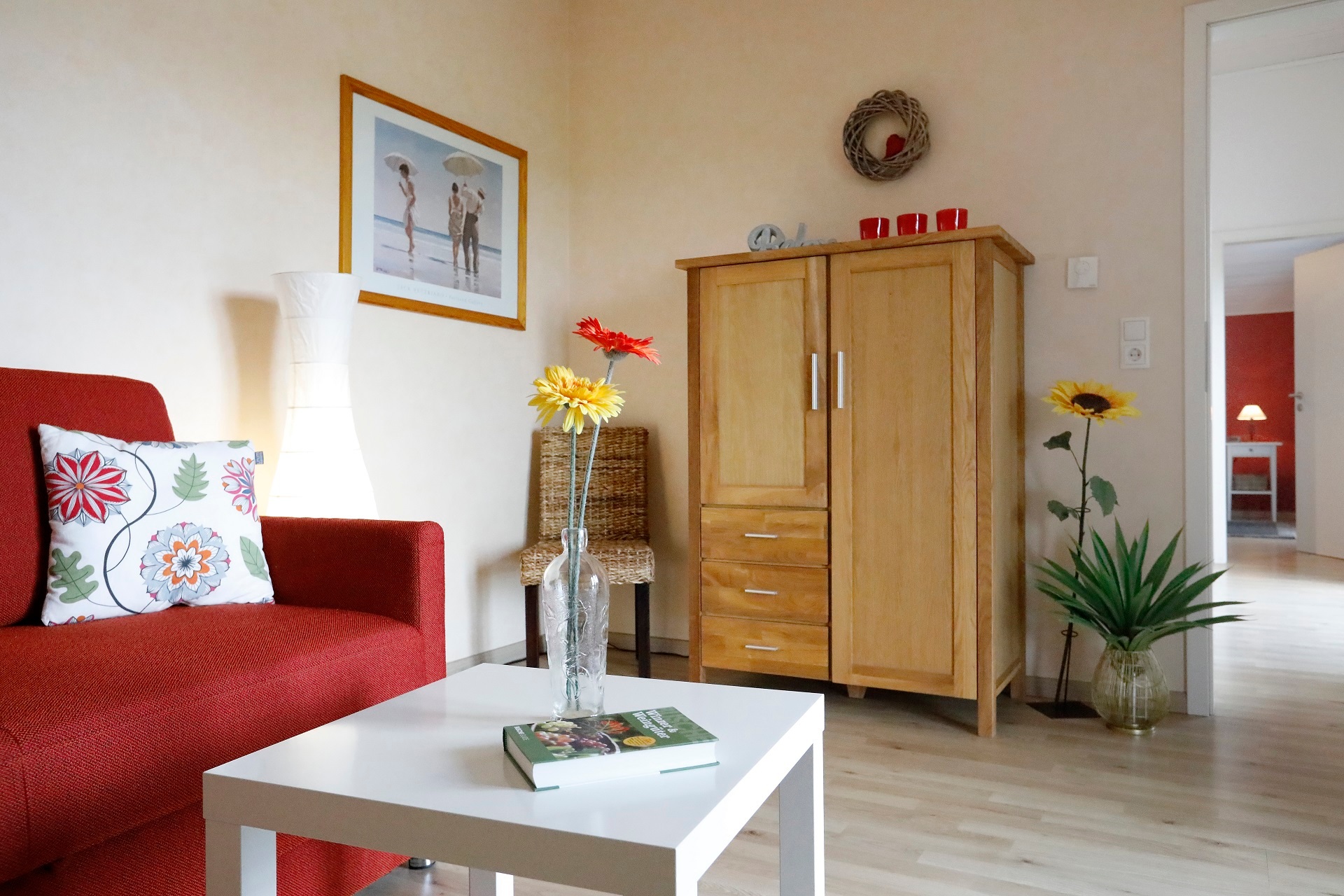 Wohnzimmer Ferienwohnung Wegner Im Vordergrund der Couchtisch der links stehenden roten Couch mit gemusterten Kissen. Im Hintergrund ein Wohnzimmerschrank und eine Kübelpflanze.