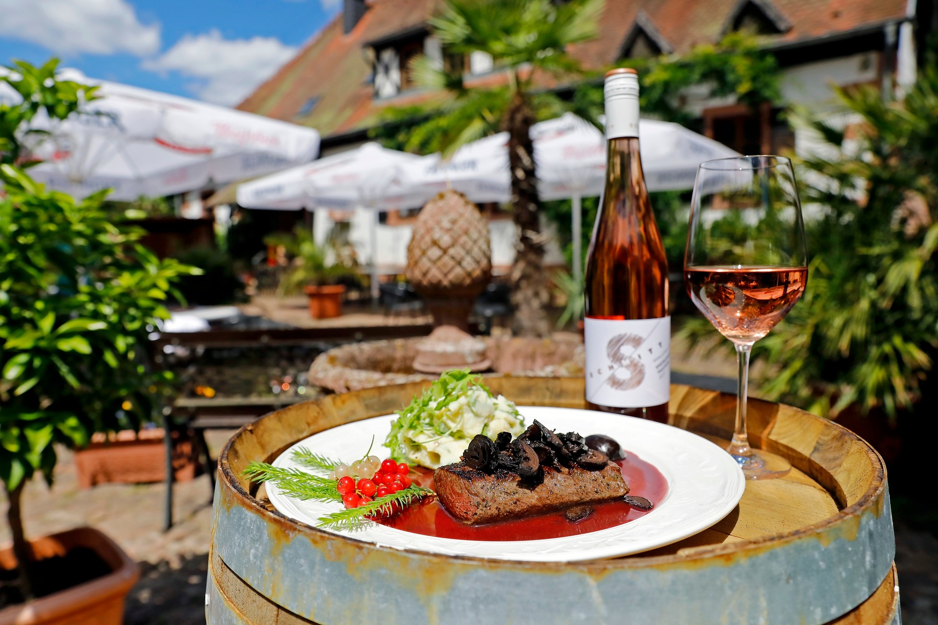 Auf einem Holzfass wurde ein Wildgericht mit dem passenden Wein angerichtet. Auf dem weißen Teller befindet sich ein saftiges Stück Fleisch mit roter Soße und Kartoffelsalat. Daneben steht ein Rose Wein des Weingut Schmitts.