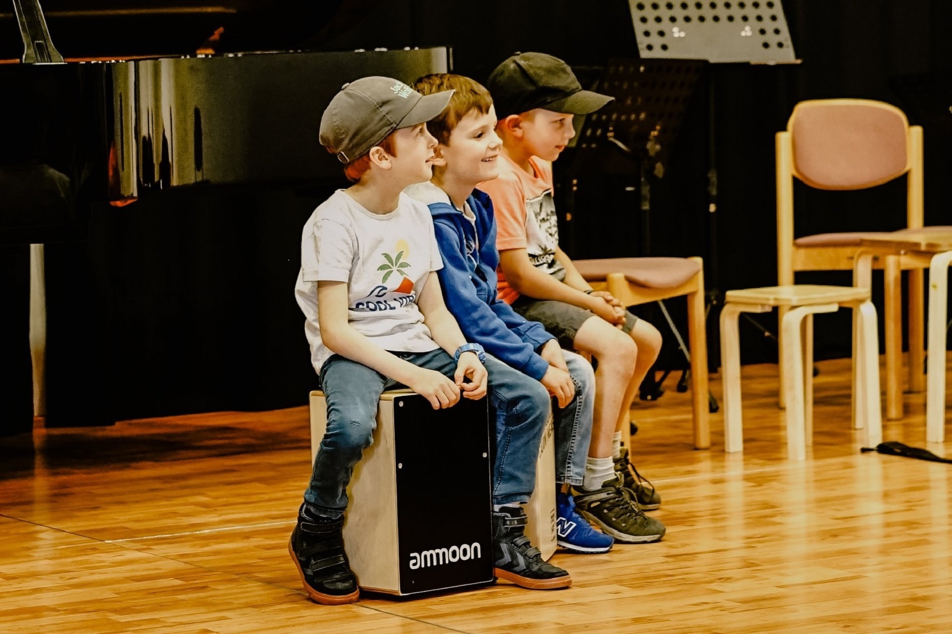Drei Jungs im Alter von ca vier Jahren sitzen auf Cahons. Sie schauen von der Kamera weg. Zwei der Kinder tragen Baseballmützen