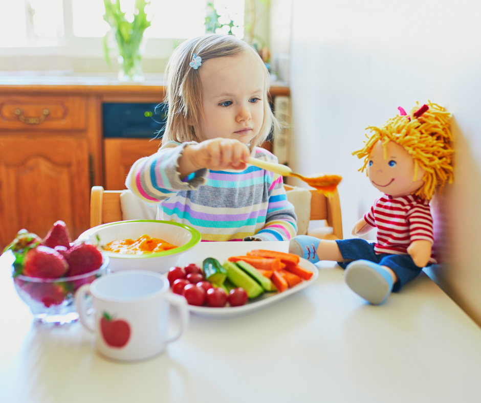 Kind sitzt am Tisch und füttert sinnbildlich eine Stoffpuppe mit Brei
