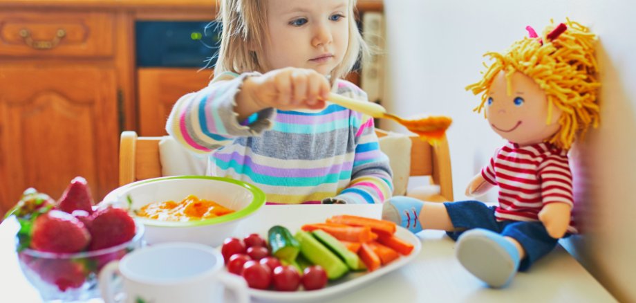 Kind sitzt am Tisch und füttert sinnbildlich eine Stoffpuppe mit Brei
